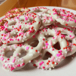 White chocolate pretzels topped with pink, red, and white sprinkles on a white plate.